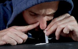 young drug addict man on hood snorting cocaine on mirror with rolled banknote at home alone